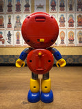 Back view of the Batten Robomaru GB-94 toy robot, showing a red circular head and torso with multiple screw holes and a battery compartment. A yellow ON/OFF switch is located on the back of the head, and a yellow knob is positioned on the torso. The arms are blue with flexible black shoulder joints, and the legs are yellow with blue feet. The design suggests electronic or interactive features, enhancing its playability and mechanical appeal.
