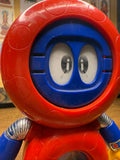 Close-up of the Batten Robomaru GB-94 figure’s torso, featuring a red circular chest with a transparent window. Inside the window is a cartoon character with a red helmet, antennae, and large round glasses, smiling cheerfully. The legs are yellow with black rectangular designs and metallic joints. This internal character detail adds charm and depth to the toy’s design, blending mechanical and animated elements.