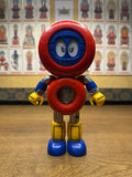 Full-body view of the Batten Robomaru GB-94 toy robot, showcasing its vibrant and stylized design. The figure has a large red circular head with a blue face and expressive eyes, a red torso with a transparent window revealing internal components, blue arms with yellow claw-shaped hands, and metallic legs with yellow and blue boots. The playful color scheme and character details highlight its appeal as a vintage children’s toy and collectible.