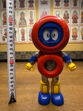 Full-body view of the Batten Robomaru GB-94 figure standing upright beside a vertical measuring tape, indicating a height of approximately 20 cm. The robot features a red circular head with blue cartoon-style eyes, a red torso with a transparent chest window, blue arms with yellow claw-like hands, and yellow legs with blue feet. The image provides a clear size reference for collectors and sellers.