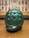 "Green sculpted object resembling a dragon egg or armored pod, featuring a metallic sheen and symmetrical overlapping scales shaped like hearts or leaves. The egg is placed on a wooden surface. In the background, a wall displays numerous small portraits of Buddhist figures, each labeled with Japanese text, with one central framed figure suggesting prominence."