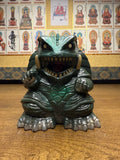 "Figurine of Gamera, the turtle-like kaiju monster, placed on a wooden surface beside a vertical measuring tape showing a height of approximately 16 cm. The creature has a fierce expression, tusks, and clawed limbs. Behind it, a wall is decorated with framed images of Buddhist figures labeled in Japanese, creating a contrast between pop culture and spiritual themes."