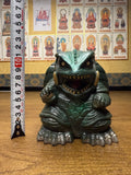 "Figurine of Gamera, the turtle-like kaiju monster, placed on a wooden surface beside a vertical measuring tape showing a height of approximately 16 cm. The creature has a fierce expression, tusks, and clawed limbs. Behind it, a wall is decorated with framed images of Buddhist figures labeled in Japanese, creating a contrast between pop culture and spiritual themes."