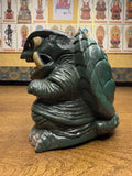 "Dark green sculpted figure of a turtle-like monster with prominent tusks, a textured shell, and a wrinkled body. The creature has a fierce expression and detailed surface textures. It is placed on a wooden surface, with a background wall displaying religious iconography of Buddhist figures in traditional attire, labeled in East Asian script."