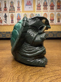 "Stylized figurine of a monstrous turtle with exaggerated features including large claws, a shell, and an open mouth filled with sharp teeth. The figure is placed on a wooden surface. The background features a wall of framed Buddhist deity images with accompanying Chinese text, suggesting a shrine or temple setting."