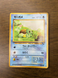 Vintage Japanese Squirtle (Zenigame) card placed on a wooden surface, showing Squirtle in a grassy landscape. The card has a blue border, 50 HP, and includes Japanese text, Water-type icon, and two attacks. Illustrated by Kagemaru Himeno.